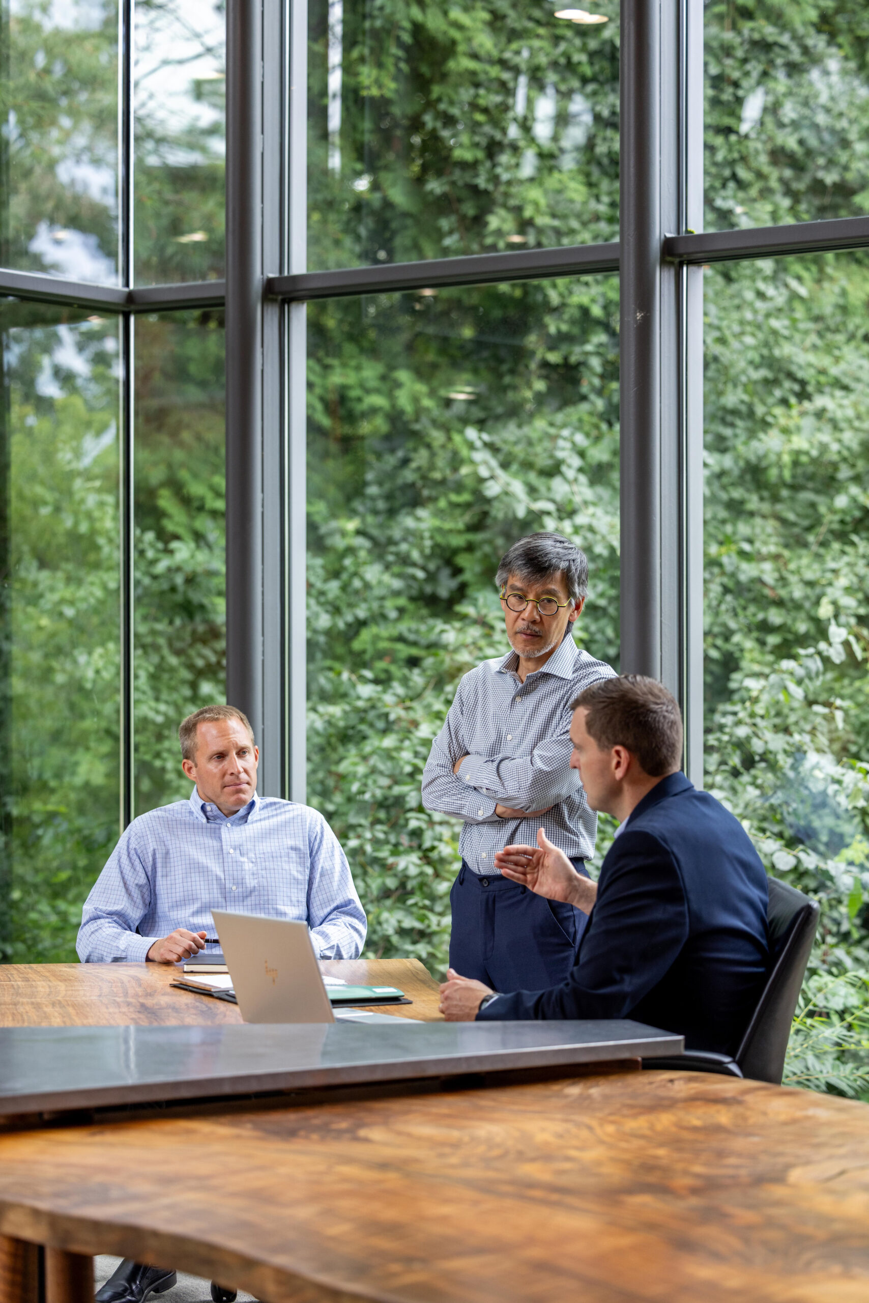 Three men in business attire have a discussion in a modern office with large windows and a view of green trees. Two are seated at a wooden table with a laptop, while one stands, listening attentively.