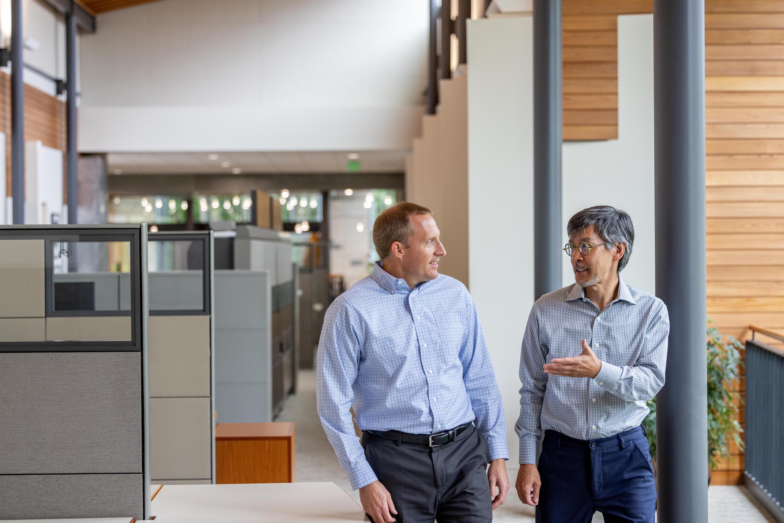 Two men in business attire walk and talk in a modern office space with cubicles, glass walls, and wooden accents. Both appear engaged in conversation, with one gesturing as they walk.